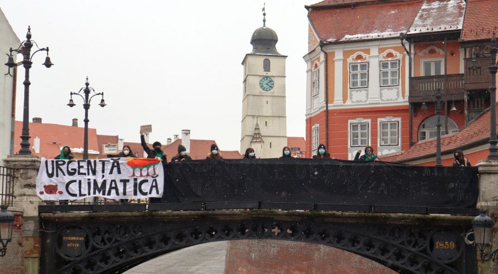 A picture of activists on a bridge, wearing masks and holding a banner reading urgenta climatica. 