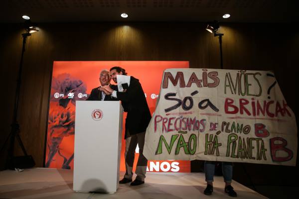 Kiko, a climate activist, interupts the Portuguese President speaking on a podium, a banner is held next to them by a further activist, with Portuguese text opposing the airport