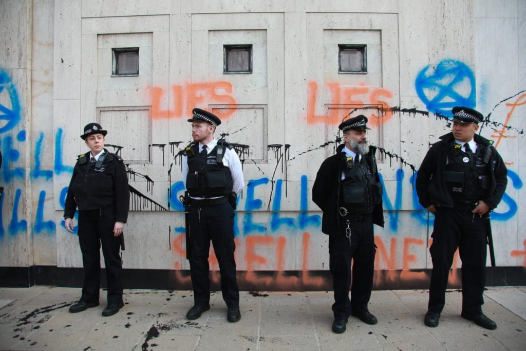 Four police officers stand in front of the Shell headquarters, graffiti covers the walls behind them. 