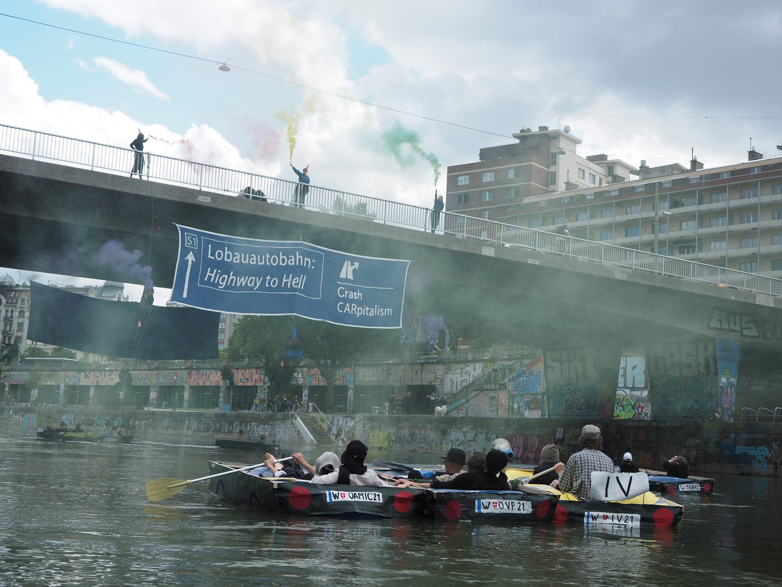 Activists in boats made to look like cars float in the Danube Canal, looking up at activists occupying the Aspern bridge with smoke.